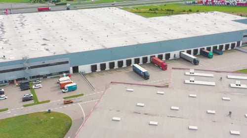 Aerial following shot of semi-trailer trucks travelling along a warehouse on the parking lot in the