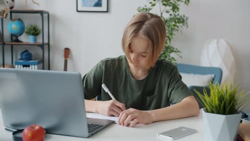 Teen Boy Writing in Notepad and Working with Laptop Doing Homework in Apartment