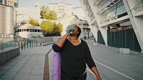 Woman Drinks Water While Walking in Urban Setting