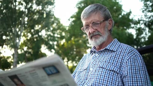 Senior in Plaid Shirt Sits on a Bench in the Park and Reads a Newspaper