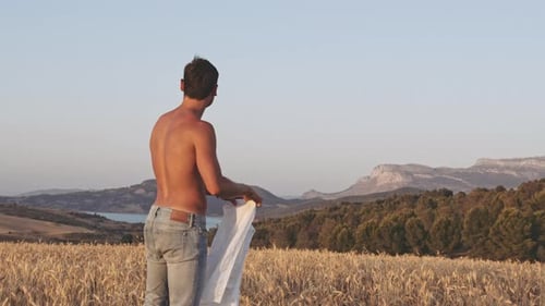 Homem joga camisa no ombro e caminha em campo aberto de trigo durante um dia ensolarado