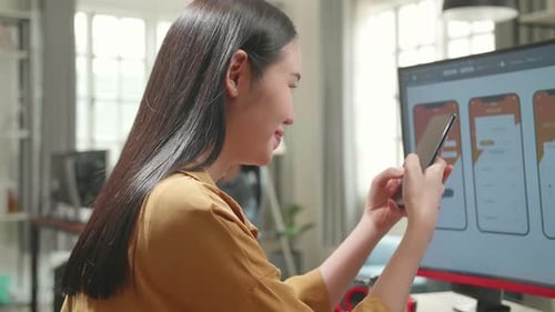 Asian Female Mobile Application Developer Works And Using Mobile Phone With Graphics On Computer