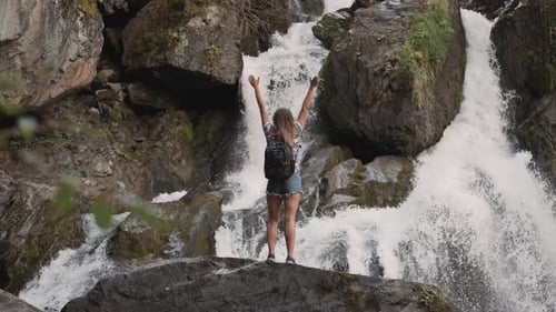 Female Tourist with a Backpack Raises His Hands Up Opposite a Large Waterfall Standing on the Edge