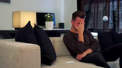 A Young, Handsome Man on a Couch Covers His Face with a Palm, Disappointed