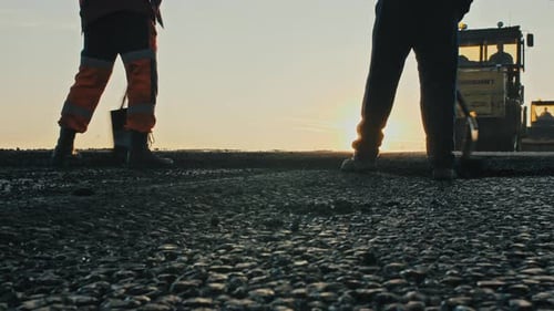 Two Road Workers in Overalls Level the Fresh Asphalt Mix Against the Background of Road Rollers