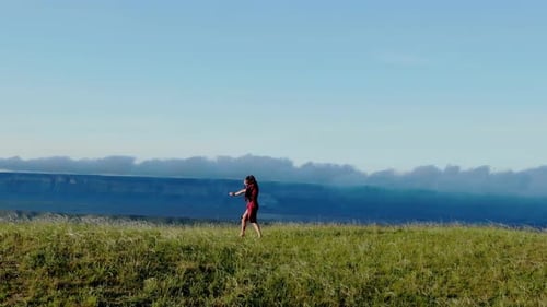 Aerial View of a Young Barefoot Woman in a Red Dress Who Dances in the Grass Against the Background