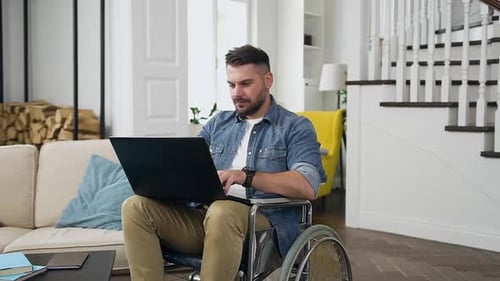 Bearded Young Man in Stylish Wear Sitting in Wheelchair and Working on Laptop at Home