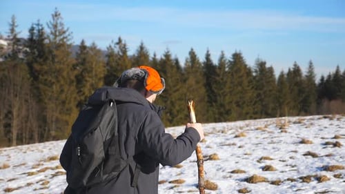 Side View of Young Hiker with Backpack and Stick in Hand Climbing on Snowy Hill in Field. Sporty Guy