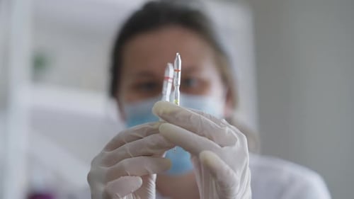 Closeup Ampoules with Liquid Drugs in Female Hands in Gloves with Blurred Caucasian Woman in Covid19