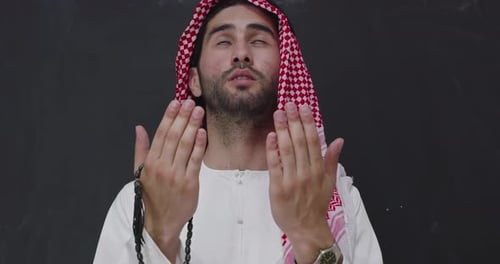 Muslimischer Mann in traditioneller Kleidung vor schwarzer Tafel betet Allah mit weit geöffneten Händen