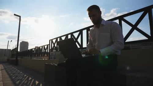 Young Employee Works on Notebook Sitting on Bench at City