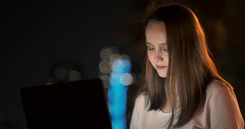 Woman Developer Sits in the Park at Night in the City and Writes Code