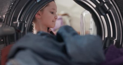 Lächelndes Mädchen in Zöpfen packt Waschmaschine aus, nimmt bunte, nasssaubere, riechende Kleidung aus