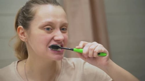 Woman in Beige Tshirt Actively Brushes Her Teeth with Green Toothbrush in the Bathroom at Home
