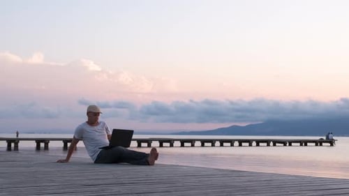 Männlicher Reisender sitzt mit Laptop am Ufer. Digitales Nomaden- oder Remote-Worker-Konzept