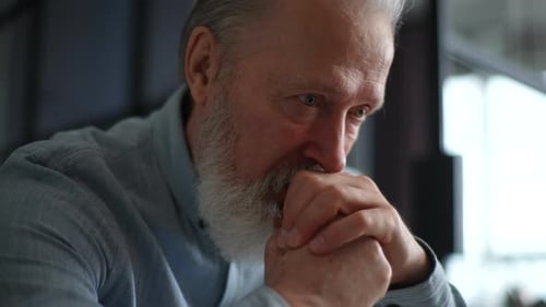 Closeup Face of Worried Unhappy Grayhaired Mature Male Put Chin on Hands Sit Indoors Feels Abandoned