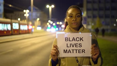 A Young Black Woman Shows a Black Lives Matter Sign to the Camera in a Street at Night