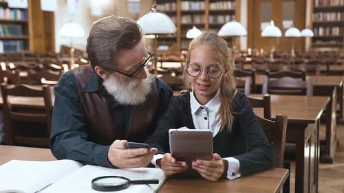 Schoolgirl Holding Ebook in Her Hands and Talking with Her Senior Bearded Grandpa in the Library