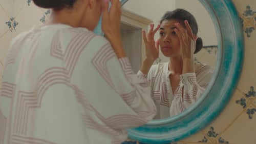 Une femme afro-américaine mécontente des années 20 touchant la peau de son visage en regardant un miroir debout dans la salle de bain