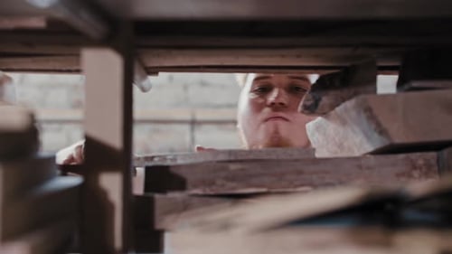 Bearded Man in a Carpentry Workshop Takes a Wooden Detail From the Shelf and Blowing Dust From It