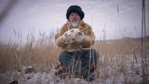 Vista frontal: xamã mongol tocando pandeiro em câmera lenta, sentado no prado nevado de inverno ao ar livre