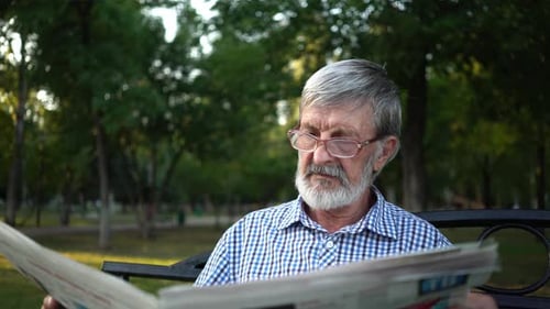 Senior in kariertem Hemd sitzt auf einer Bank im Park und liest eine Zeitung