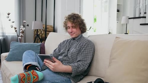 Man Dressed in Casual Clohes Sitting on the Cozy Sofa in Contemporary Apartment and Using Tablet PC