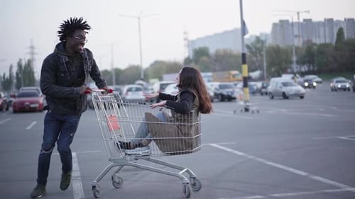 Cheerful Young African American Boyfriend and Caucasian Girlfriend Having Fun Rolling Shopping Cart