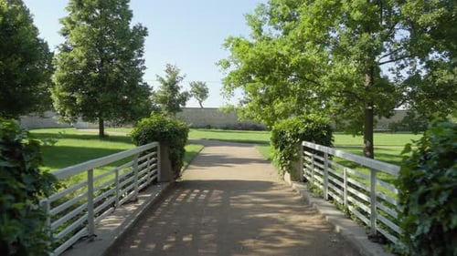 A Pathway in a Green Park with Trees and Bushes Over a Bridge with White Fence
