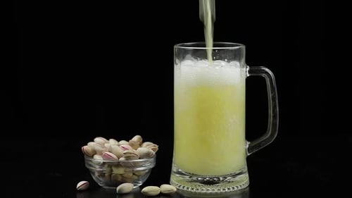 Beer Is Pouring Into Glass on Black Background. Bowl of Pistachios Nuts
