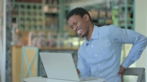 Young African Man with Back Pain Using Laptop in Cafe