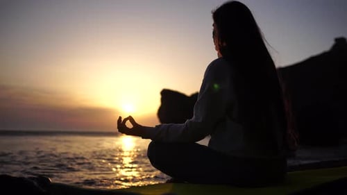Young Woman with Long Hair Practicing Stretching Outdoors on Yoga Mat By the Sea on a Warm Sunset