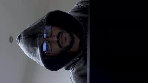 A vertical video of a hacker working on his laptop computer wearing a dark hoodie covering his head,