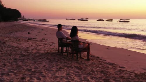 Young couple relaxing on calm exotic beach, sitting on chairs over white sand, watching beautiful su