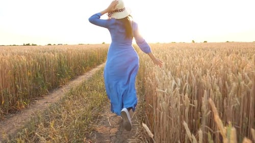 Beautiful Woman in a Blue Dress and Hat Runs Through a Wheat Field at Sunset