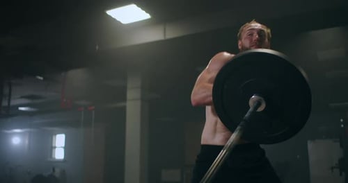 The Boy Pushes the Barbell in the Gym with One Hand. An Athlete Without a T-shirt in a Dark Room