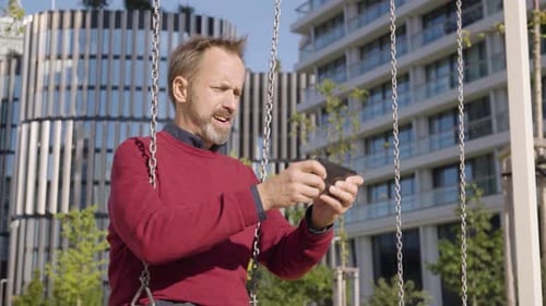 Man Using Phone on Swing in Urban Park