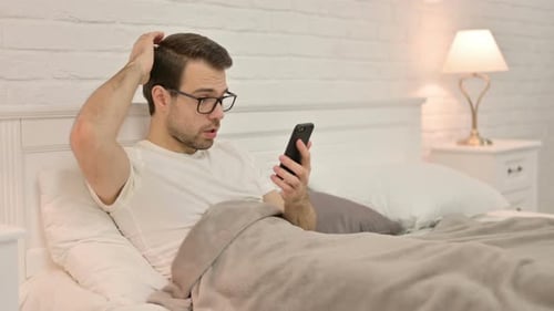 Man in Bed Using Mobile Phone, Looking Surprised