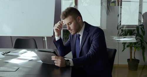 Selbstbewusster, fokussierter Geschäftsmann trägt Anzug, sitzt im Büro am Tisch und nutzt digitales Tablet für die Arbeit