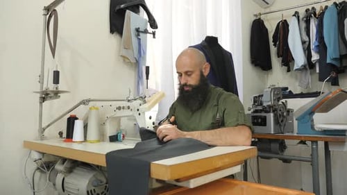 Bearded Tailor Sewing Dark Fabric at Machine