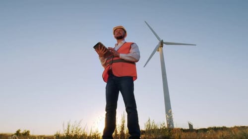 The Engineer a Man in a Helmet and Vest Looks at a Digital Tablet and Looks Away Into the Distance
