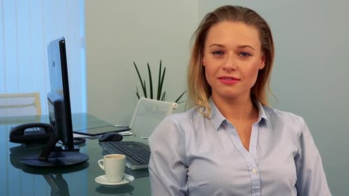 A Young, Attractive Woman Sits at a Table in an Office, Smiles and Points at the Camera