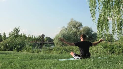 Young man meditating doing yoga exercise by riverside in park in morning