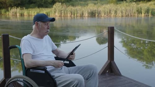 Senior Man Using Tablet on Lake Dock in Wheelchair