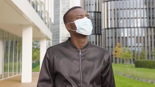 A Young Black Man in a Face Mask Thinks About Something - Office Buildings in the Blurry Background