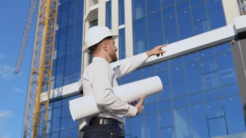 An Engineer in a White Shirt and Helmet Works on the Construction of a Modern Glass Building and