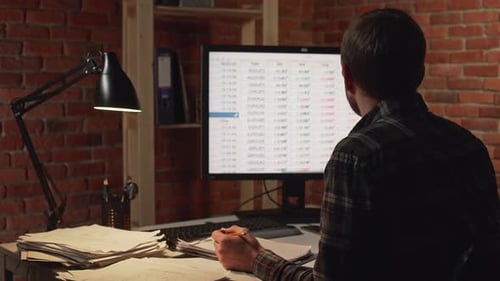 Back View of Caucasian Male in Glasses Sitting in Front of Monitor and Making Notes in Papers in