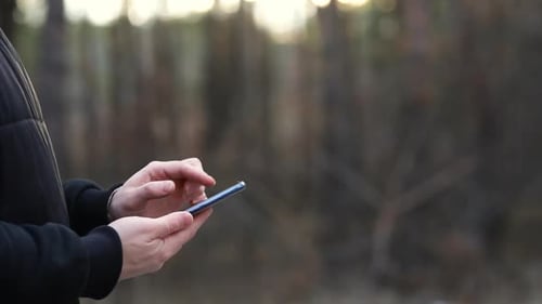 The guy is typing on the phone in front of forest