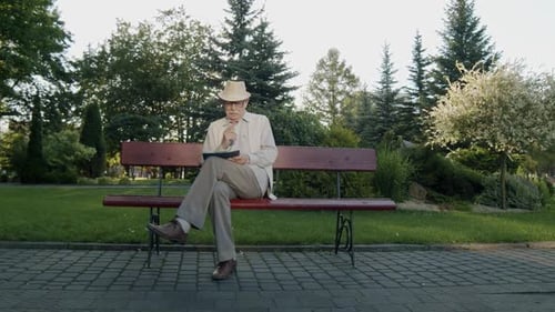 Thoughtful Senior Man Sitting on Park Bench and Noting on Papers in Summer Day