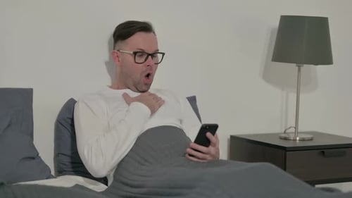Man in Bed Reacts to Phone with Surprise